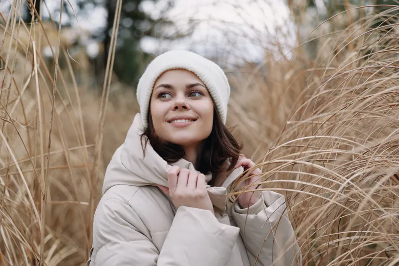 Woman in a beige coat and white knit hat stands among tall grasses outdoors, smiling softly, capturing a serene winter portrait with natural light, cozy fashion vibe and gentle movement in airy