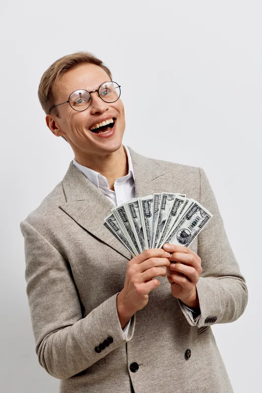 Smiling young man with glasses wearing a light suit holding US dollar bills in hand, expressing happiness and success, isolated on plain white background, studio portrait, business achievement concept