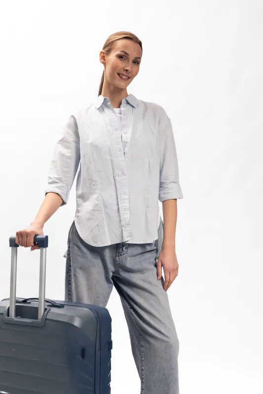 Young woman in casual outfit stands with a suitcase on white background. Travel concept with relaxed female traveler ready for a trip and smiling confidently to camera.