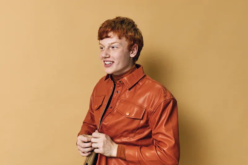 Young man with red hair and freckles wearing an orange leather jacket standing against beige background looking sideways with thoughtful expression. Casual fashion style portrait.