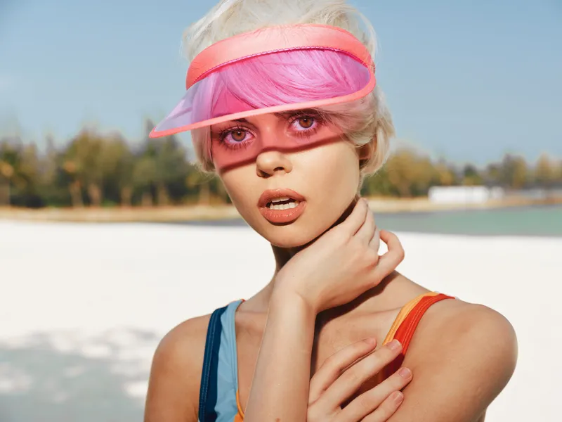Portrait of a young woman wearing a pink visor on a sunny beach, posed with a hand at her neck, featuring bright colors, bold summer style, and a striking fashion moment.