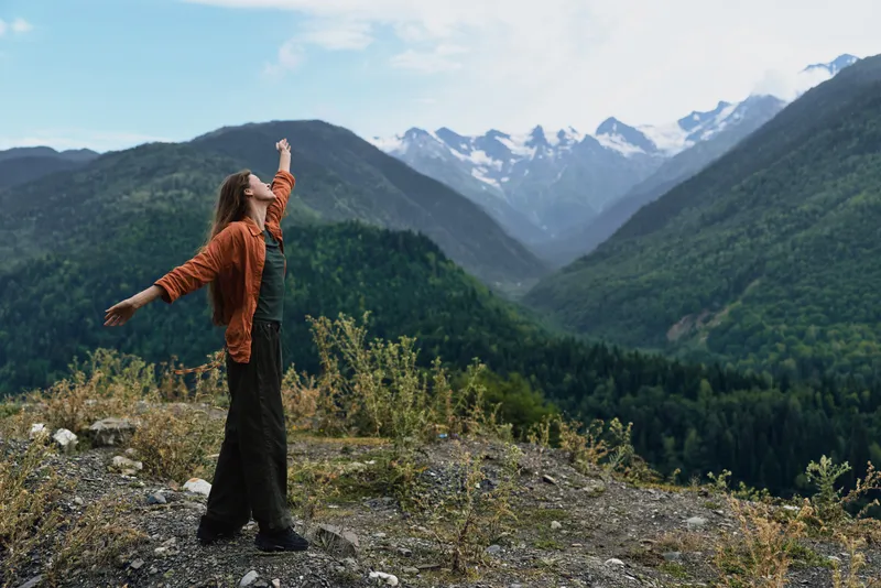 person mountains nature freedom authenticity inclusivity outdoors landscape adventure, woman on rocky ridge with arms raised facing distant snowcapped peaks, serene exploration and mindful moment