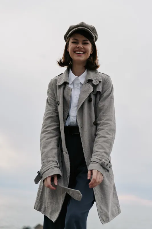 Smiling young woman in textured grey coat and matching hat stands outdoors against cloudy sky. Casual fashion, autumn outfit, and happy mood concept with natural light and relaxed pose.