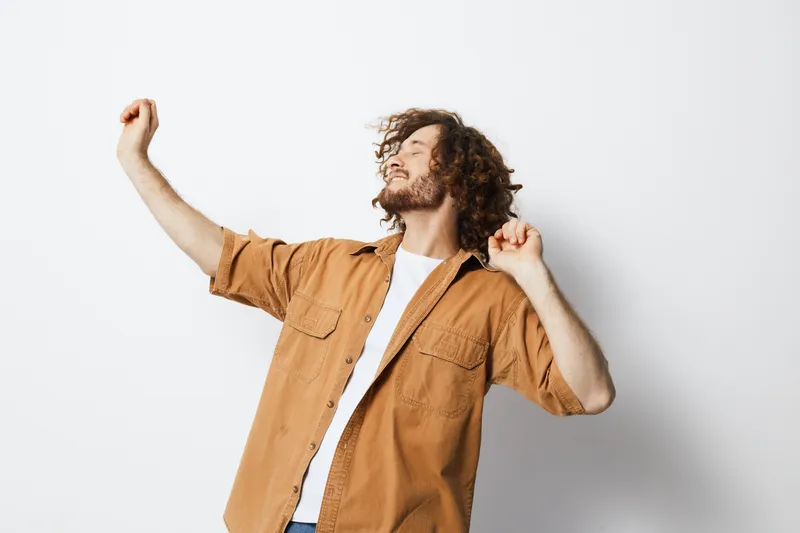 Young man enjoying music, dancing joyfully against a light background, showcasing a vibrant lifestyle and expressive emotions in a casual outfit