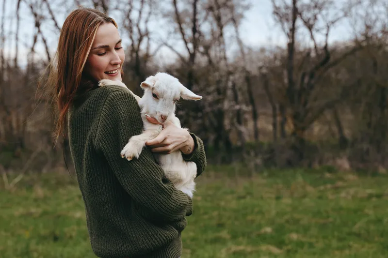 Smiling woman holding a goat kid outdoors in a green field, conveying lifestyle and affection. Warm, natural lighting highlights happiness and tenderness in a rustic countryside scene.