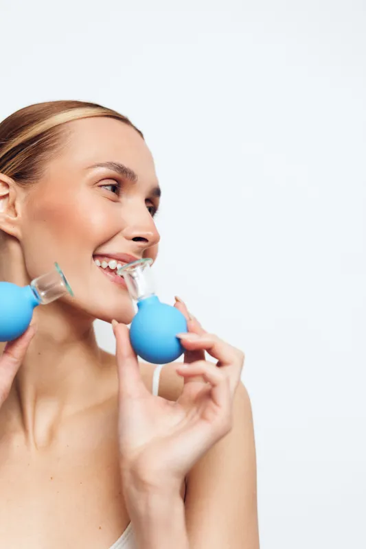 Young woman with clear skin applies facial cupping therapy using blue silicone cups for skin care and anti aging treatment isolated on white background.