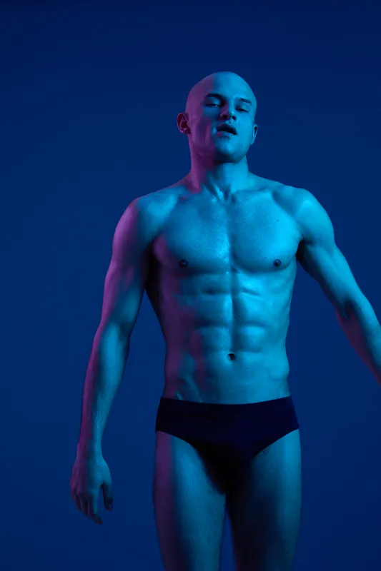 Athletic man posing gracefully under blue lighting, showcasing strength and confidence with a dynamic posture. A striking visual representation of male beauty.