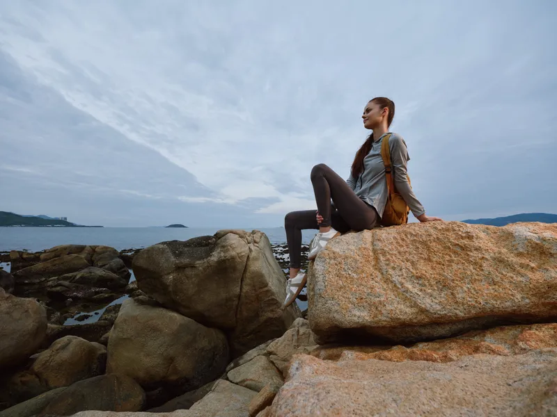 Young woman enjoying nature on rocky shore, wearing casual attire, with serene expression under a cloudy sky, reflecting a connection to the environment