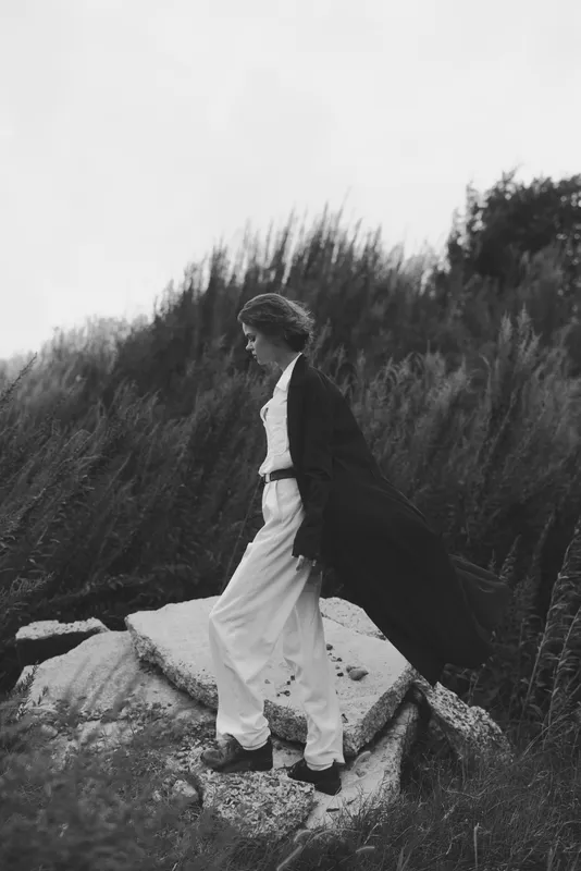 Fashion, outdoor, coat, silhouette, monochrome, wind, shoreline, journey, movement, moody scene of a figure in a long coat walking along a rocky coast amid grasses and sea breeze