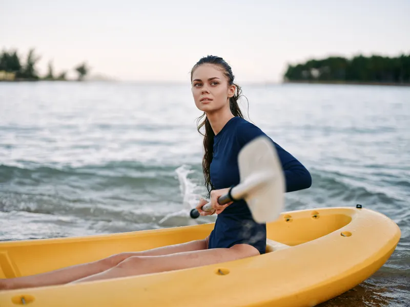 Female kayaker sits in a bright yellow kayak on calm coastal water, wearing a dark wetsuit and gripping a paddle. She gazes ahead with calm focus.