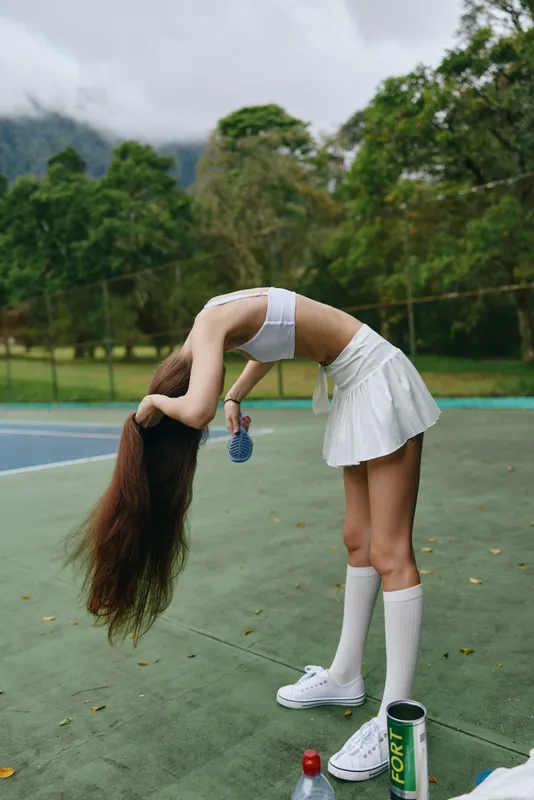 Young woman in a sporty outfit brushing long hair on a tennis court The scene showcases her white crop top and skirt, combining athleticism with style, surrounded by greenery