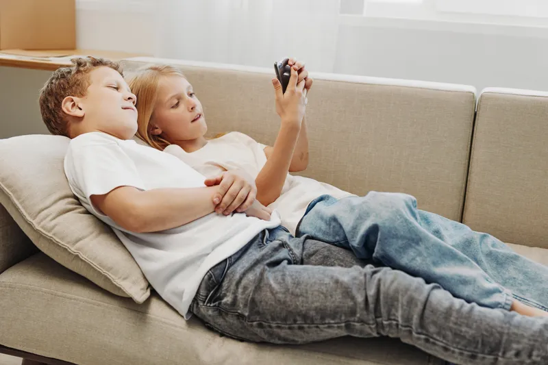 Two relaxed children lying on a beige sofa, watching something on a smartphone together. Casual clothing and natural light create a cozy, calm, and intimate home atmosphere.
