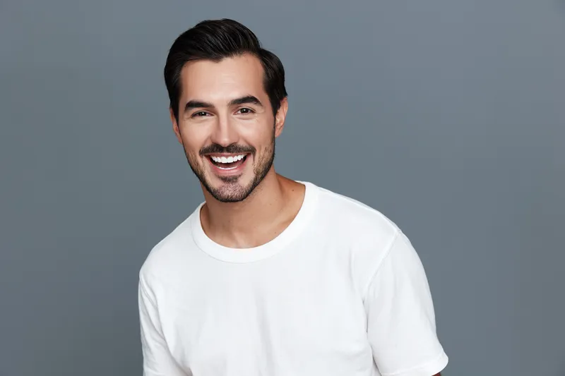 Smiling man with dark hair wearing a white t-shirt, standing against a light gray background, showcasing warmth and approachability in a cheerful pose.
