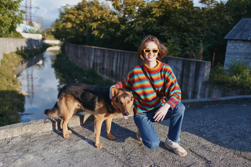 Woman in her early 20s kneeling by a canal with a German shepherd, smiling and wearing a colorful striped sweater, yellow sunglasses, crossbody bag and casual jeans outfit.