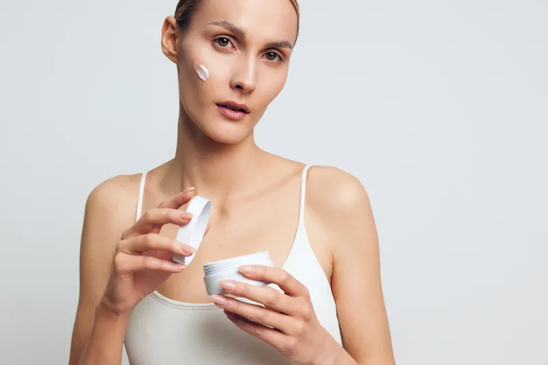 Young woman applying face cream on cheek, skincare routine, natural look, soft lighting, neutral background, casual outfit, focused expression, beauty care concept