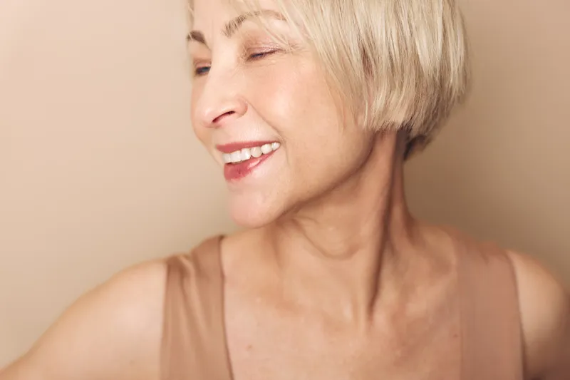 Smiling mature woman with short blonde hair looks away with closed eyes, expressing joy and confidence on beige background. Concept for happiness, aging, and natural beauty.