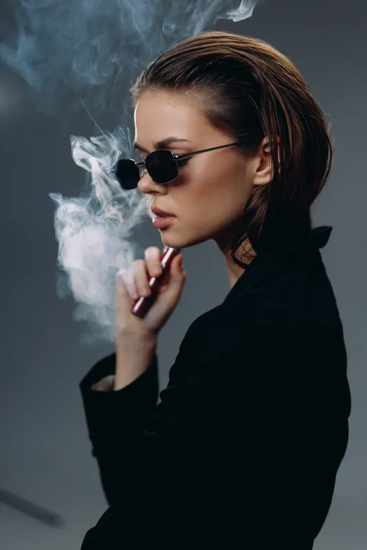 Stylish woman smoking electronic cigarette with smoke in dark background wearing sunglasses and black outfit.