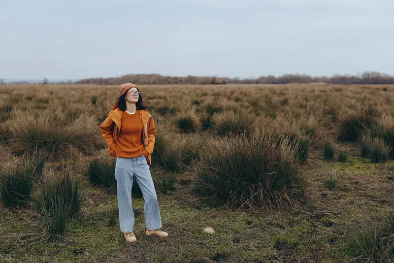 Woman lifestyle outdoors in a field of grass standing with relaxed pose wearing orange sweater and light jeans daylight creates calm wellness mood in countryside landscape during spring