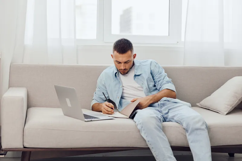 Thoughtful man sitting on a couch, writing notes in a cozy living room. Relaxed atmosphere with natural light, showcasing creativity and personal growth.