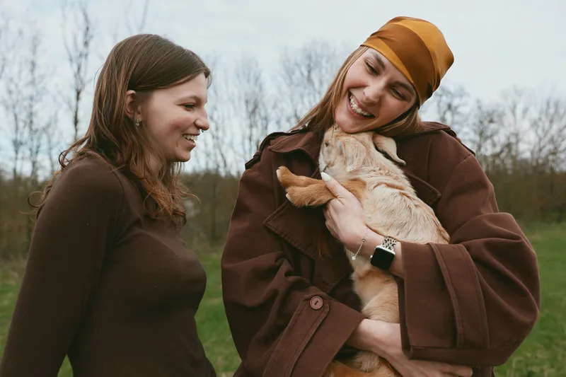 Two women outdoors with a goat kid, smiling and warm, sharing a gentle moment in a meadow, showcasing friendship, lifestyle, and animal companionship under natural light
