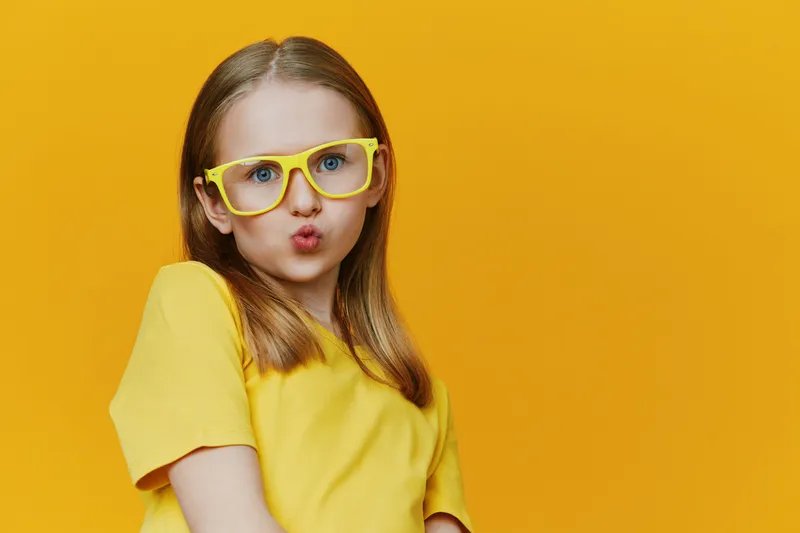 A cheerful young girl with yellow glasses and a yellow t shirt poses playfully in front of a bright yellow background, radiating joy and confidence