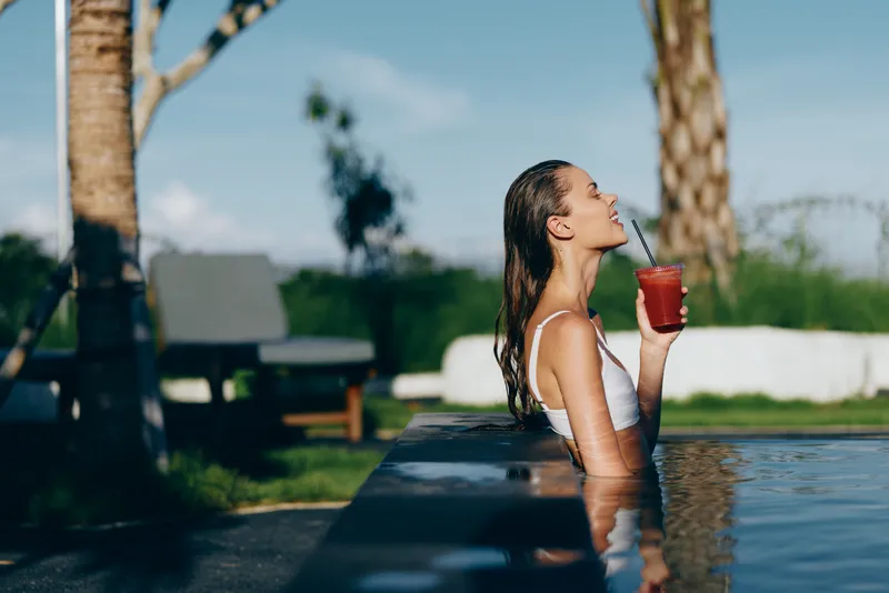 Cheerful woman in a poolside setting, drinking a bright juice with a straw, sunlit water reflections, relaxed posture, tropical vibes, vibrant colors, calm afternoon moment, lifestyle