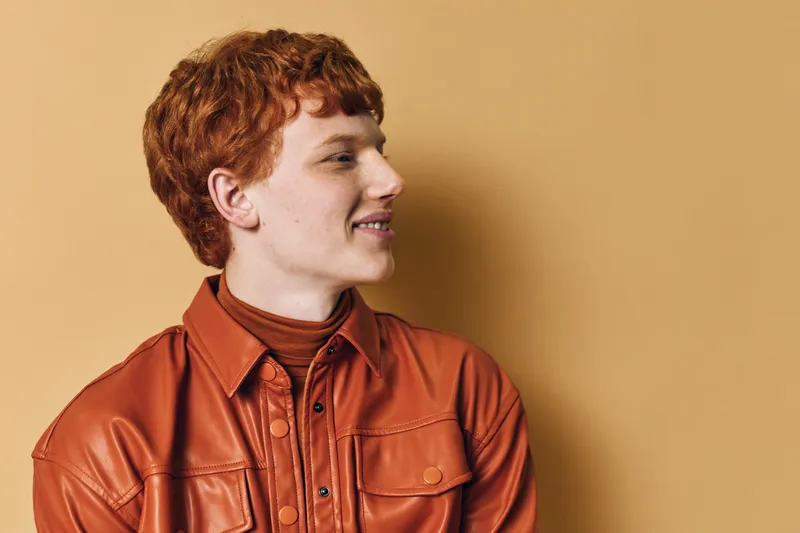 Young man with short red hair and freckles smiling while wearing a brown leather jacket and turtleneck against a beige background in studio portrait.