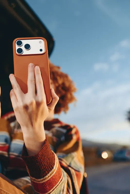 Woman holds a smartphone for a selfie outdoors, framed against a blurred street and travel road, casual portrait lifestyle moment in warm sweater, sunny sky, candid hands close