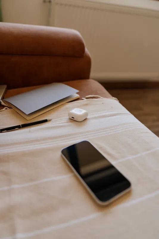 Casual home scene with a smartphone, notebook, pen and wireless earbuds on a striped table beside a couch, suggesting a relaxed productive woman lifestyle at home