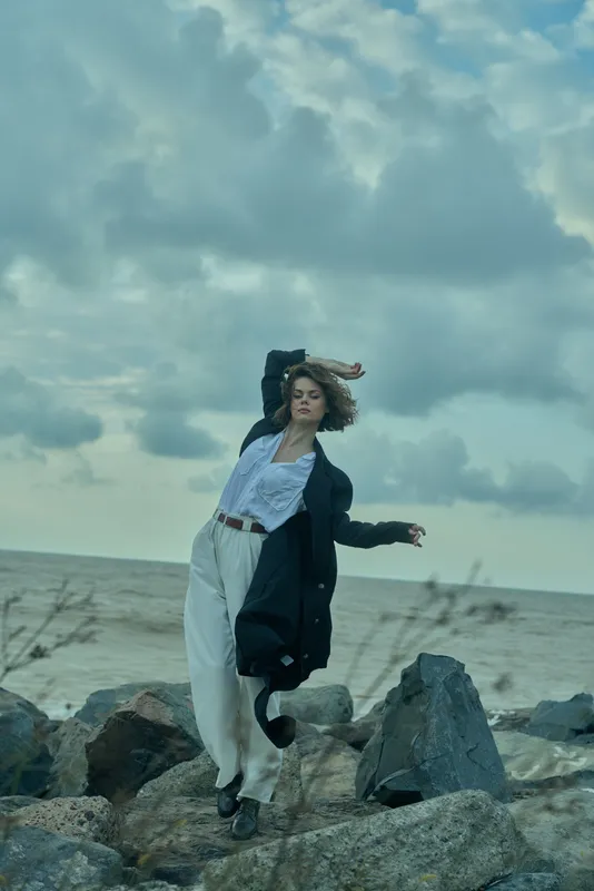 Woman standing on jagged rocks by the sea, wind catching hair, wearing light blouse, wide pants and dark coat, dynamic motion against a cloudy coastal sky, moody fashion moment
