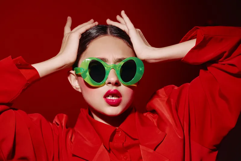 Fashionable woman in bright red outfit wears oversized green sunglasses with shiny glossy lips standing against vibrant red background. Creative beauty editorial concept.
