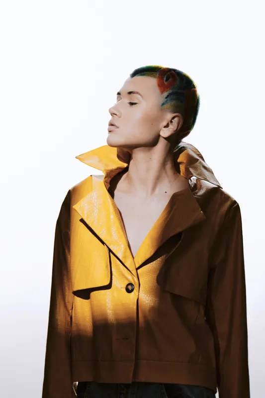 woman with colorful shaved hair wearing a yellow leather jacket poses with confidence against a white background in studio lighting