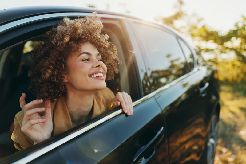 A young woman with curly hair leans out of a moving car window, smiling as warm light filters through the trees on a sunny road trip.
