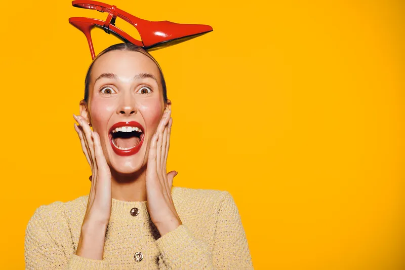 Excited woman with bright red lipstick and beige sweater holds face with hands, wearing red high heel shoe on head. Vibrant yellow background creates playful and energetic mood.