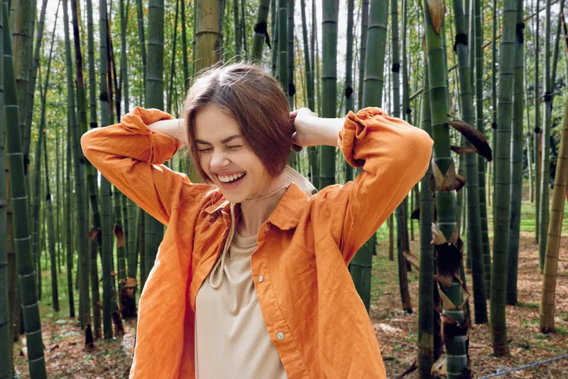 Woman smile bamboo forest outdoors nature portrait jacket, happy candid moment with eyes closed, casual orange outerwear during a serene leisure walk through a green bamboo grove.