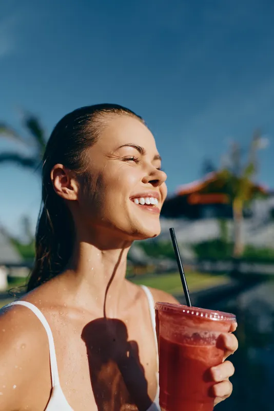 Sunny woman enjoys a refreshing juice outdoors, smiling softly, relaxed summer mood, white top, bright natural light, palm trees behind, close up portrait, lifestyle moment, carefree vacation vibe