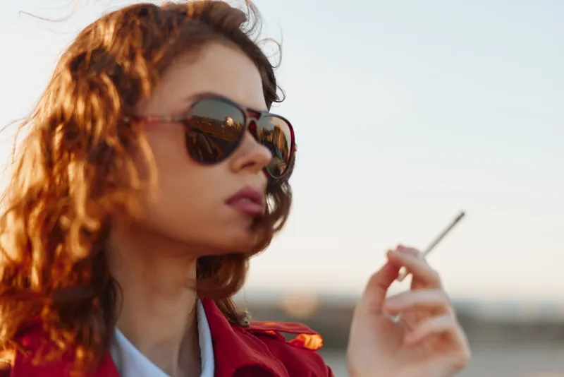 A young woman with red hair and sunglasses exhales smoke while posing in bright daylight, conveying a moment of rebellion and confident attitude.