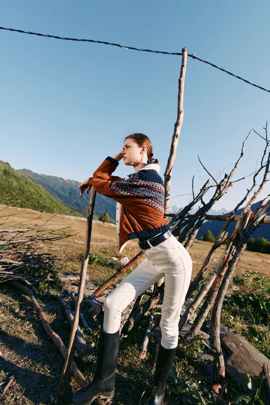 Woman in sweater and boots leaning on rustic fence in countryside mountains, portrait of outdoor lifestyle, hiking fashion and rural scenery with clear blue sky and grass.