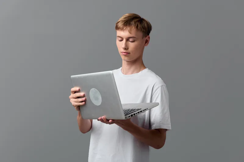 Young man holding a laptop, focused on the device screen, casual workspace with minimal backdrop, learning and working with technology, exploring design and code, daily productivity moment, screen