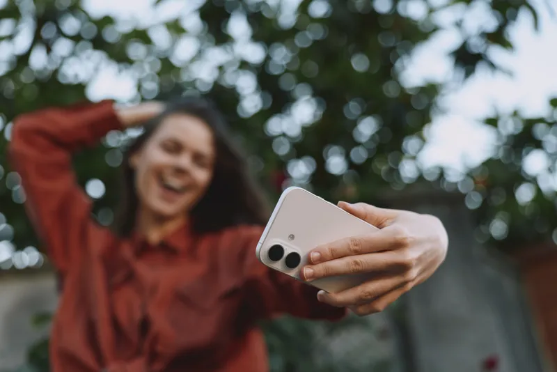 Smiling woman taking a selfie outdoors, holding a smartphone toward the camera, wearing a red top with a leafy background. Casual lifestyle moment, cheerful mood, self expression