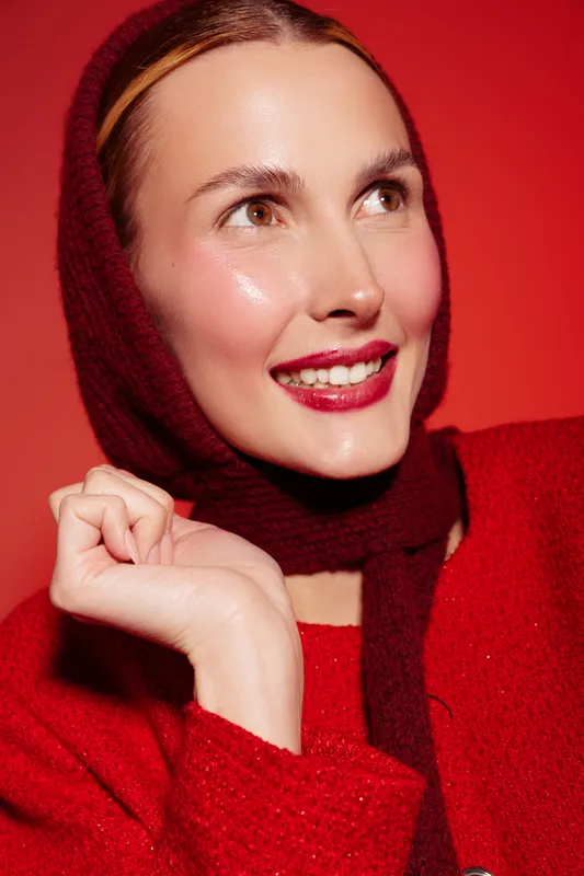 Smiling woman with bright red lipstick wears a red sweater and matching scarf on red background. Close-up portrait with glowing skin and joyful expression. Concept of beauty and fashion.