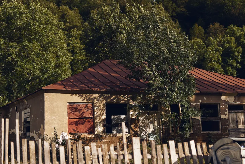 Rustic countryside house with weathered walls, red roof tiles, and a wooden fence framed by lush trees in a tranquil rural setting
