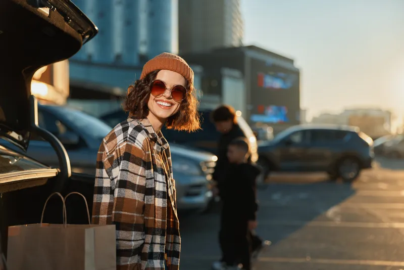 Woman smiling after shopping at parking lot in city sunset, candid lifestyle portrait with authenticity, golden hour glow, mindful living and emotional storytelling atmosphere.