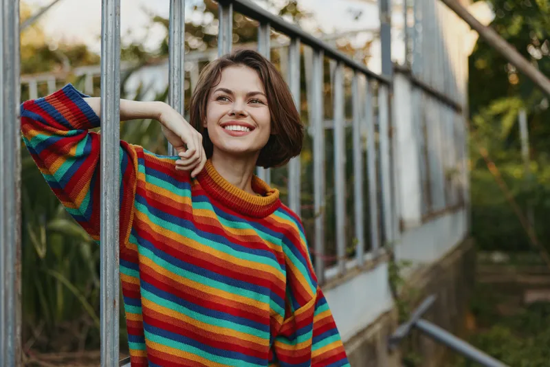 Woman in colorful striped sweater leaning on a greenhouse frame, smiling and looking up with short brown hair, casual outfit, sunny outdoor lifestyle portrait for fashion or travel.