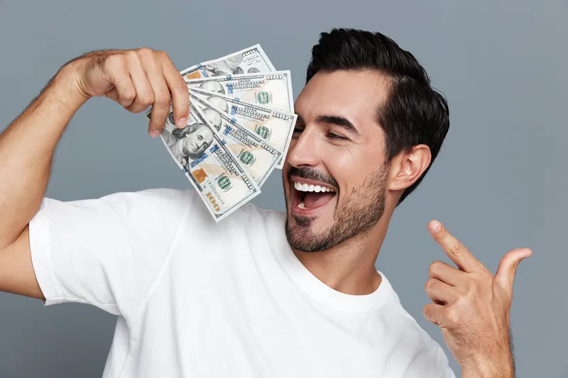 Happy smiling young man holding cash, showing enthusiasm and excitement. Isolated on a plain solid gray background, capturing a joyful financial moment.