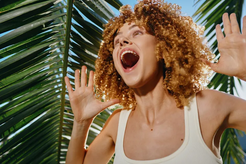Happy woman with curly hair enjoying sunny tropical environment, surrounded by green palm leaves and bright blue sky, expressing joy and freedom with open mouth and raised hands.