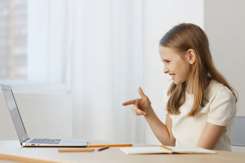 Young girl engaging in a virtual call with a laptop, showing joy and enthusiasm while sitting at a desk in a bright room, ideal for educational content