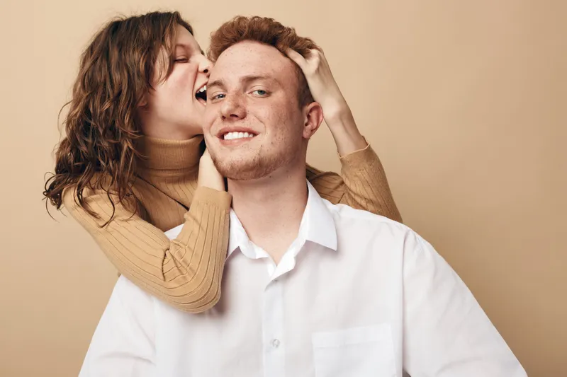 Happy young couple in casual clothes share playful moment, woman hugging man from behind and smiling. Intimate relationship, love, joy, and affection concept on beige background.