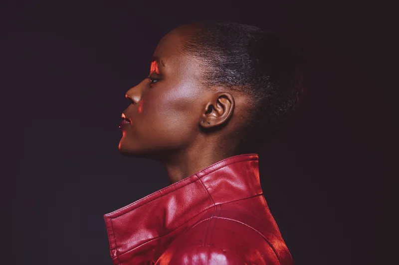 Profile of a confident dark skinned African woman wearing a red leather jacket against a black background. Portrait shows strong features and stylish attitude in studio lighting.