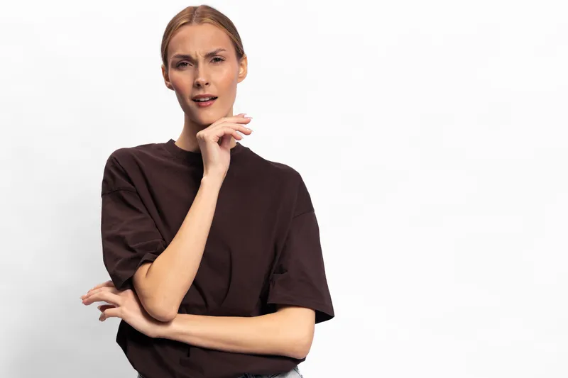 Confused young woman in dark t shirt looks puzzled with raised eyebrows and hand near face, isolated on white background. Concept of doubt, uncertainty, and questioning expression.